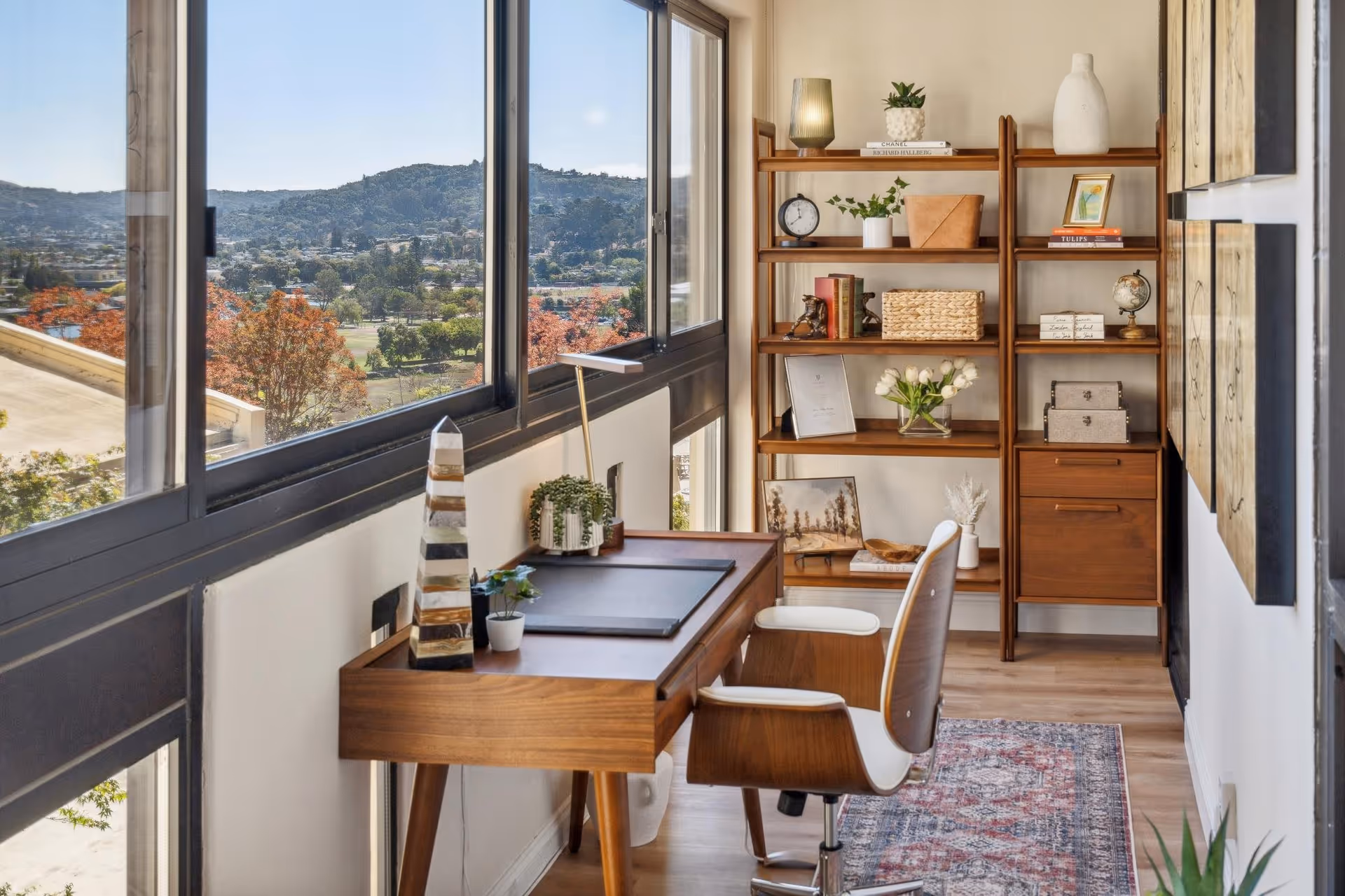 A bright and cozy office space with large windows offering a scenic view of hills and trees. The room features a wooden desk with a modern chair, a desk lamp, and small potted plants. In the background, there is a wooden shelving unit decorated with books, plants, a clock, and decorative items. A patterned rug covers part of the wooden floor.