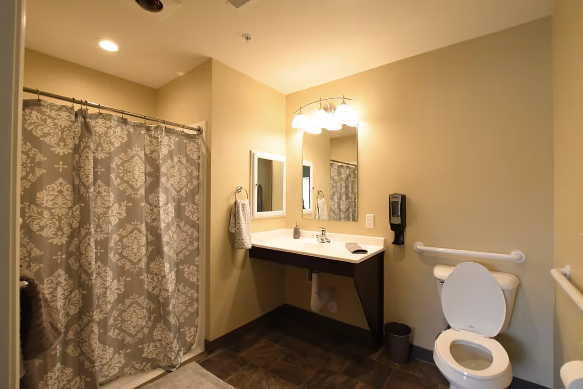 A bathroom with a shower area covered by a patterned shower curtain, a wall-mounted sink with a mirror and light fixtures above it, a towel hanging on a ring, a soap dispenser, and a toilet with grab bars on the walls for accessibility.