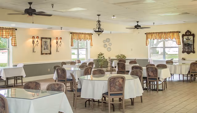 A bright dining room with multiple round and rectangular tables covered with white tablecloths. Each table is surrounded by wooden chairs with patterned cushions. The room has large windows with checkered valance curtains, wall sconces, ceiling fans, and decorative plants and wall art.