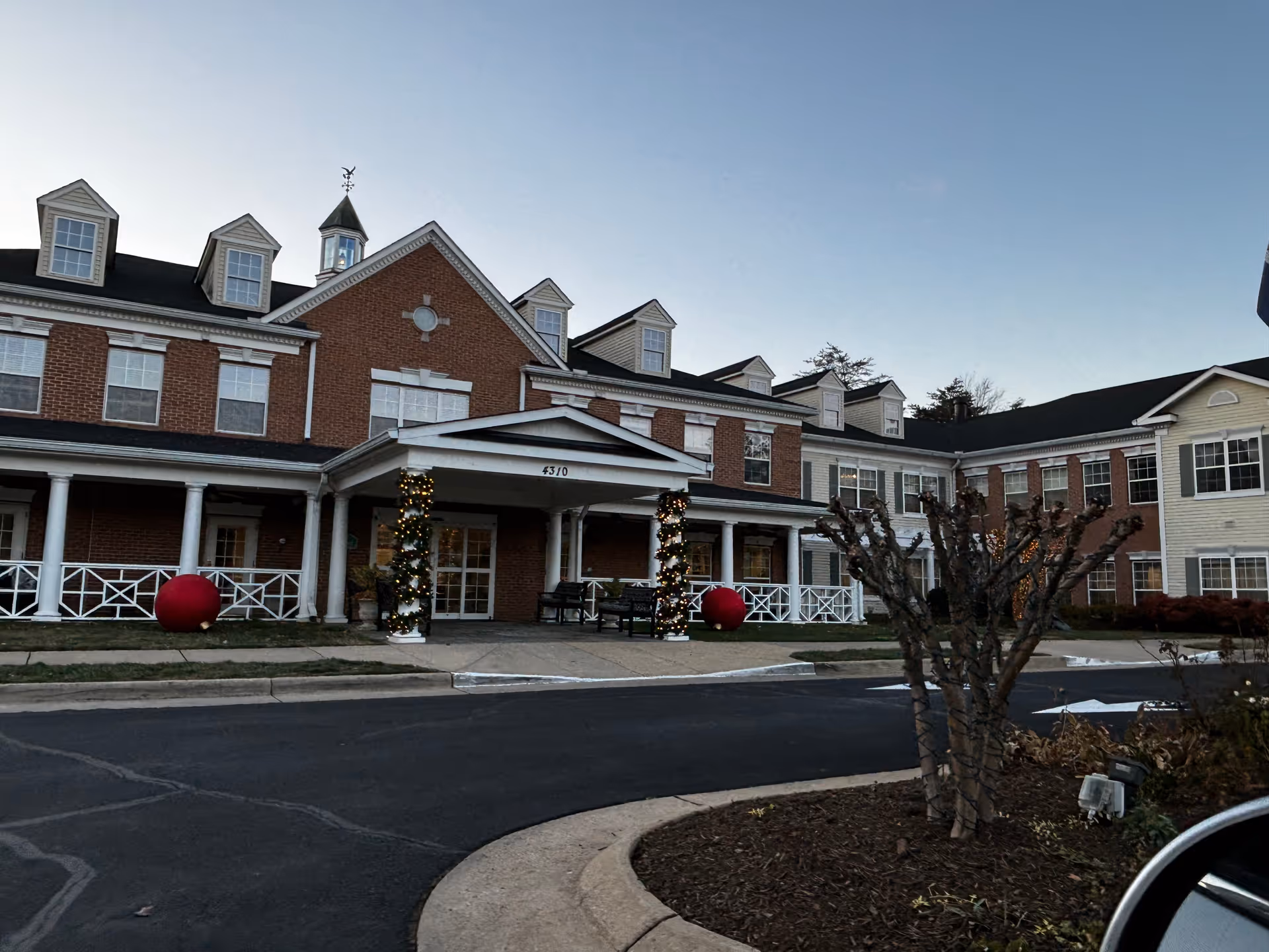 Exterior view of a large senior living facility building with brick and light-colored siding, multiple windows, and a covered entrance decorated with string lights and large red ornaments. There is a curved driveway and some landscaping with pruned bushes in front.