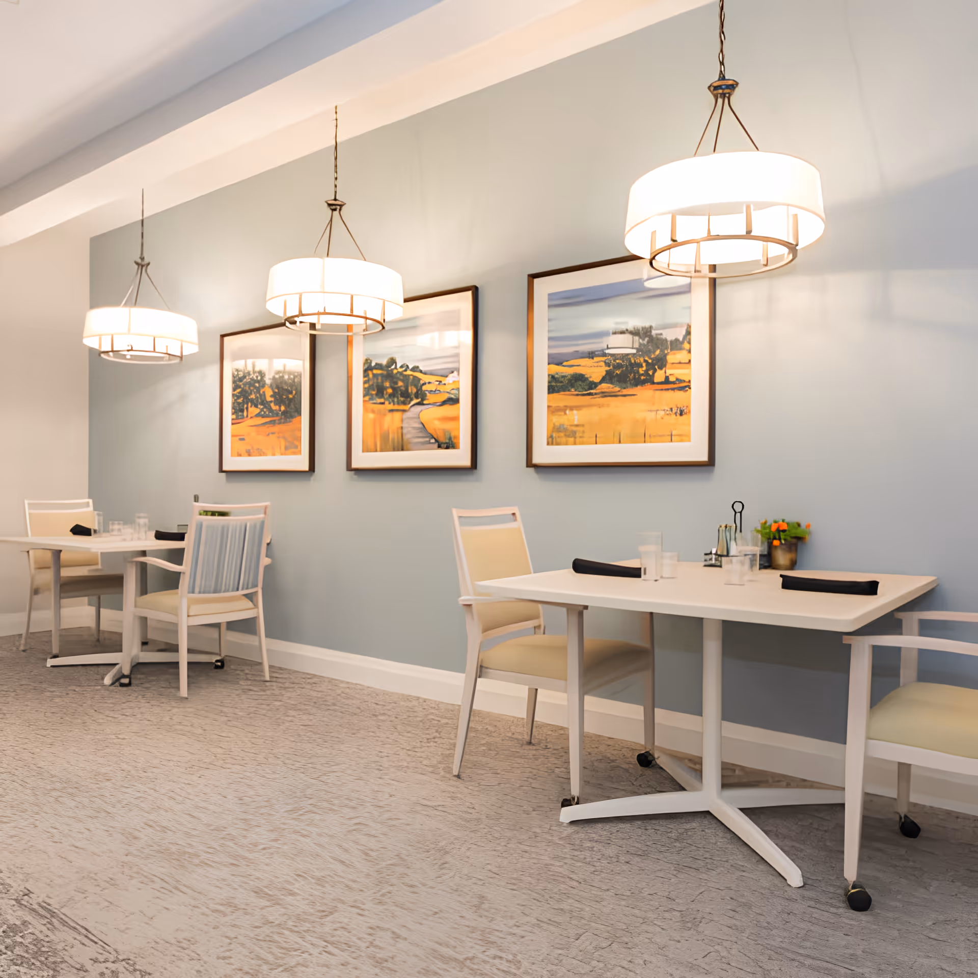 Light-filled dining area with small tables and chairs, pendant lights, and framed landscape paintings on a pale blue wall.