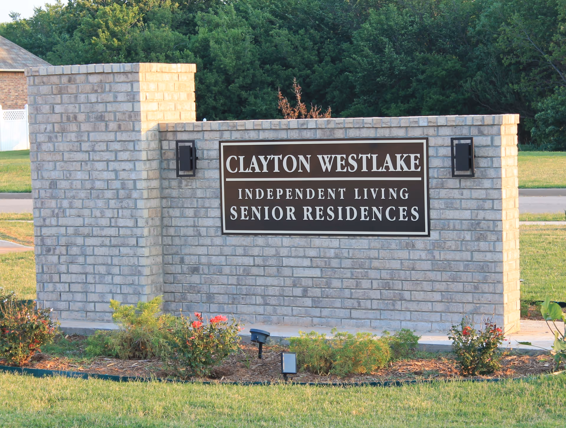 A brick sign for Clayton Westlake Independent Living Senior Residences, surrounded by grass and small plants, with trees in the background.