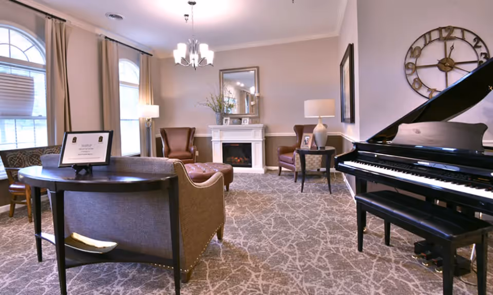 A cozy living room area with a grand piano on the right, two brown leather armchairs, a white fireplace with a mirror above it, a side table with a lamp, and a round table with a framed sign. The room has large windows with beige curtains and patterned carpet flooring.