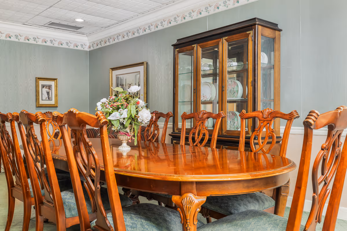 Formal dining room with a long polished wooden table, matching chairs, a floral centerpiece, and a china cabinet against the wall.