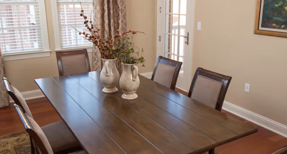A dining room with a rectangular wooden table surrounded by six chairs with cushioned seats and backs. Two decorative vases with artificial plants are placed on the table. The room has hardwood flooring, beige walls, a window with blinds and curtains, and a glass door leading outside.