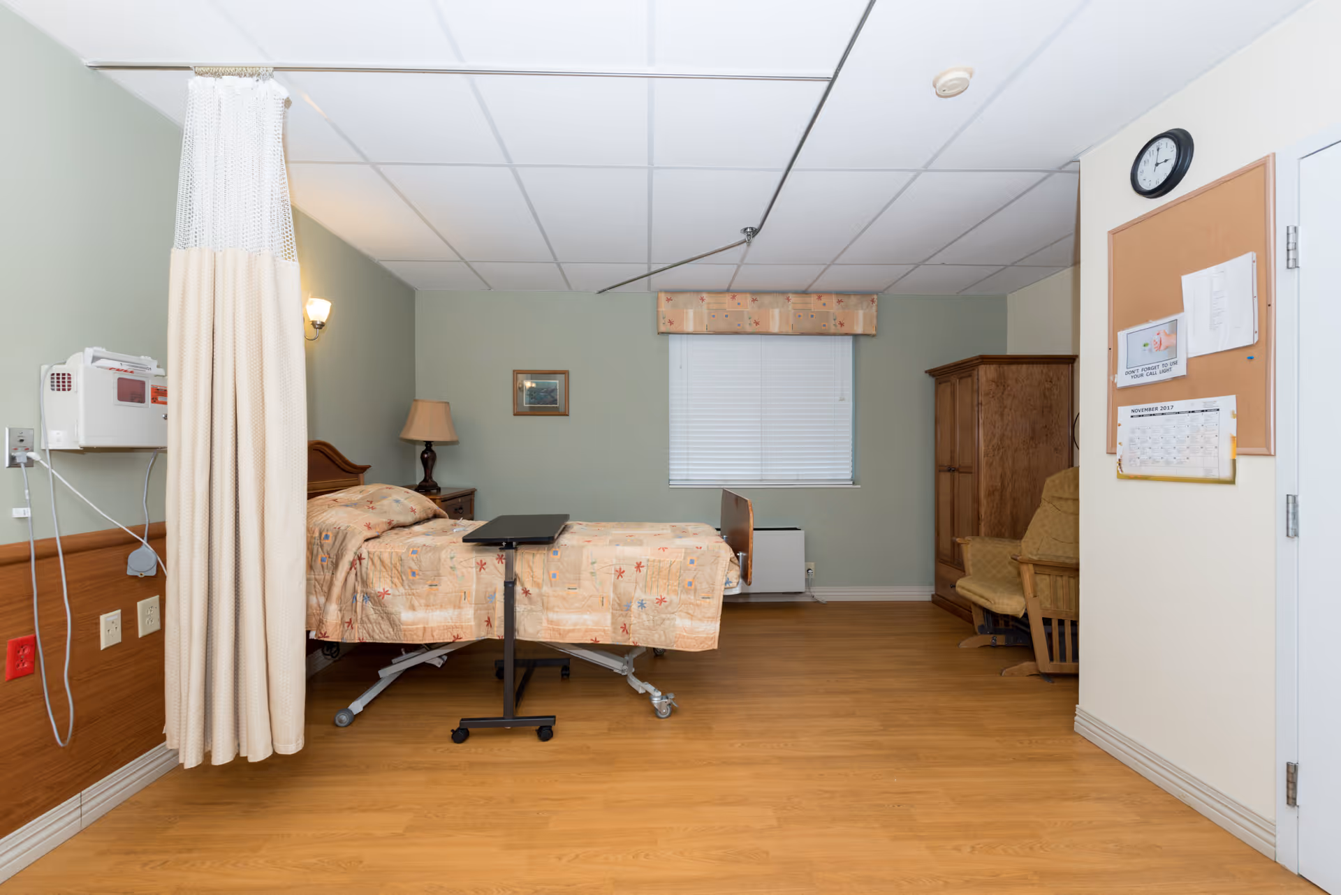 A rehabilitation facility room with a hospital bed covered in a patterned bedspread, a bedside table with a lamp, a privacy curtain partially drawn, a wooden armoire, a cushioned chair, a cork bulletin board with papers and a calendar, and a wall clock above the door. The room has light green walls and wood flooring.