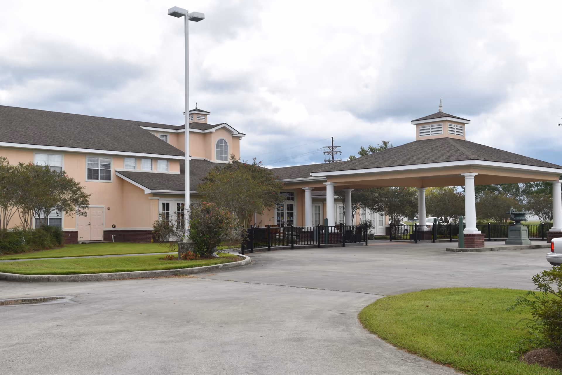 Exterior view of a senior living facility with a covered drop-off area supported by white columns. The building is light peach with white trim and has multiple windows. There is a paved driveway and some green landscaping with trees and grass around the building. The sky is cloudy.