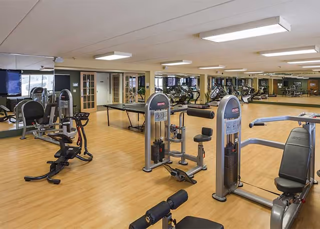 A spacious fitness room with various exercise machines including stationary bikes and weight machines, a ping pong table in the background, large mirrors on the walls, and wooden flooring.