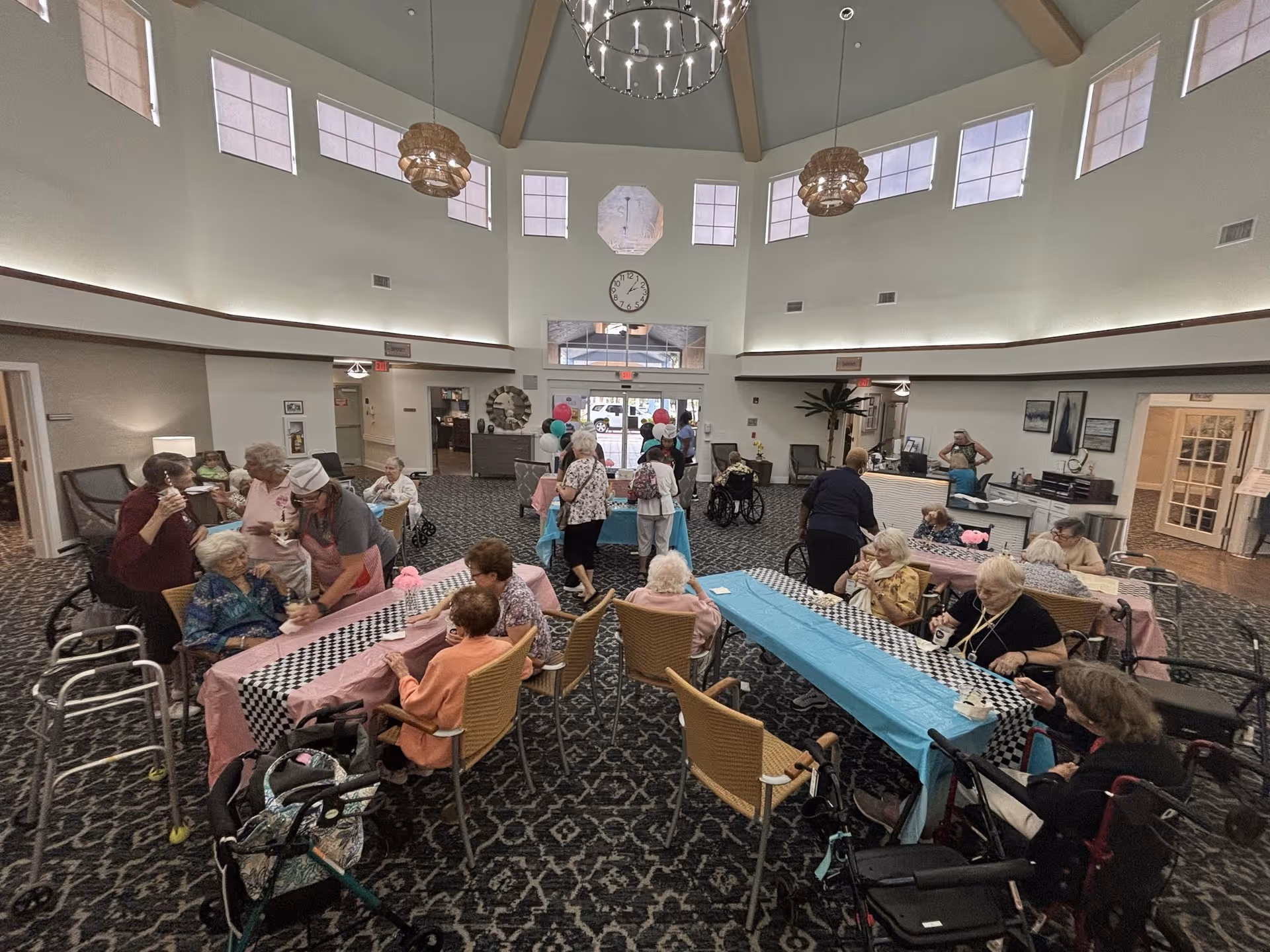 A spacious common area in a senior living facility with high ceilings and multiple windows near the ceiling. Several elderly residents are seated around tables covered with pink and blue tablecloths, engaging in activities and socializing. Some residents are using walkers and wheelchairs. Staff members are assisting and interacting with the residents. The room has patterned carpet flooring and a clock on the wall above the entrance.