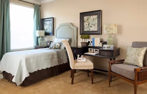 A cozy bedroom featuring a bed with a light-colored quilt and decorative pillows, a window with green curtains, a desk with a chair, two table lamps, framed floral artwork on the wall, and an armchair with a patterned cushion.