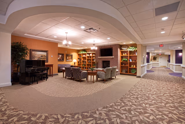 A spacious and well-lit common area in a senior living facility featuring a seating arrangement with armchairs and a coffee table centered on a carpeted floor. The room includes a brick fireplace with a mounted flat-screen TV above it, flanked by wooden bookshelves decorated with plants and ornaments. A black piano is positioned against one wall, and framed artwork hangs on the walls. The area opens into a hallway with additional lighting and decor.
