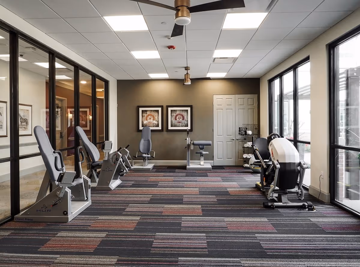 A fitness room with exercise machines including chest press and leg curl equipment. The room has large windows on the right side allowing natural light, a ceiling fan, framed artwork on the back wall, and a patterned carpet floor.