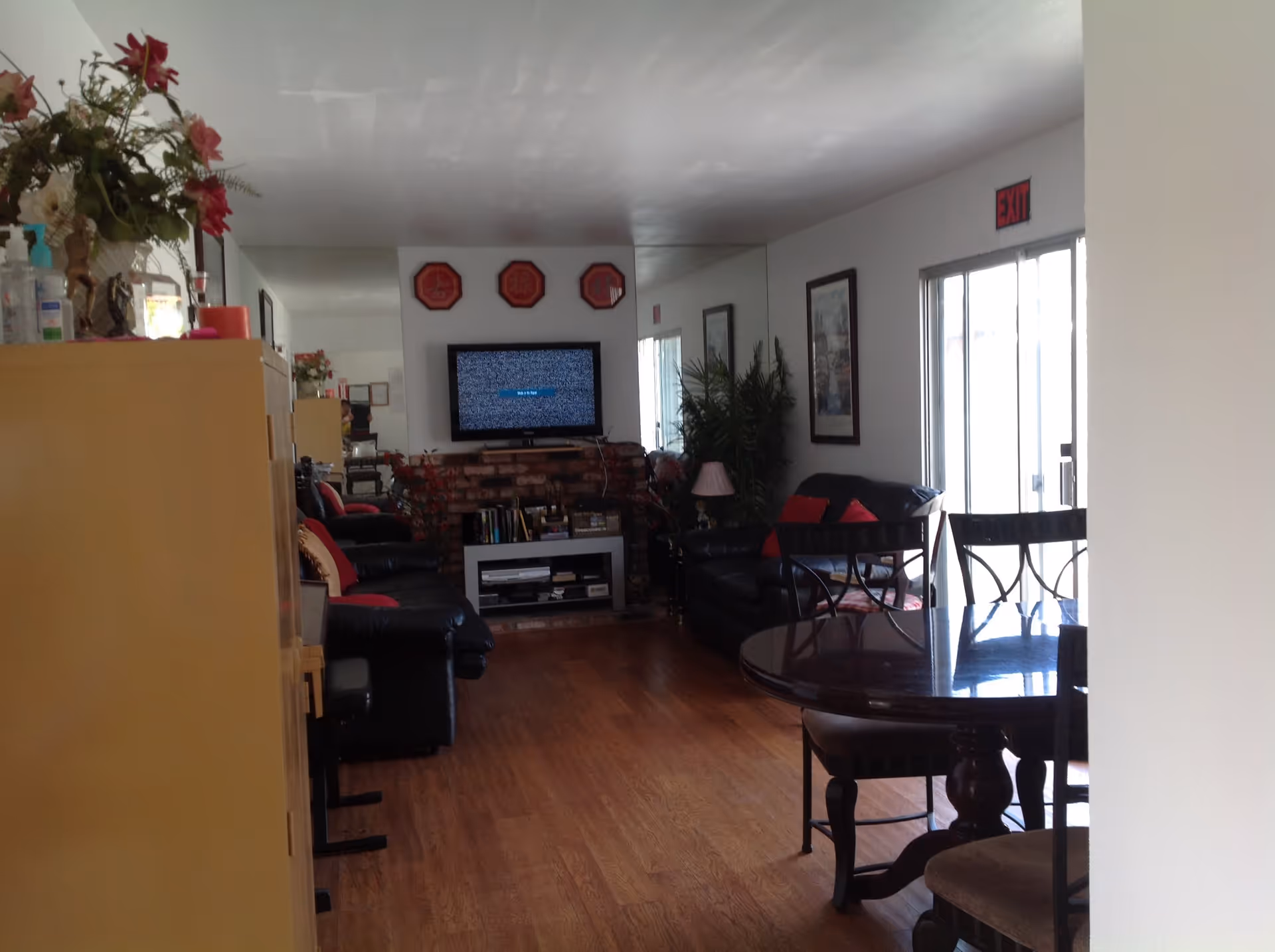 Interior view of a living room area with wooden flooring, black leather sofas with red cushions, a round wooden dining table with chairs, a TV mounted on a brick wall, decorative plants, and framed pictures on the walls. There is an exit door with a red exit sign above it.