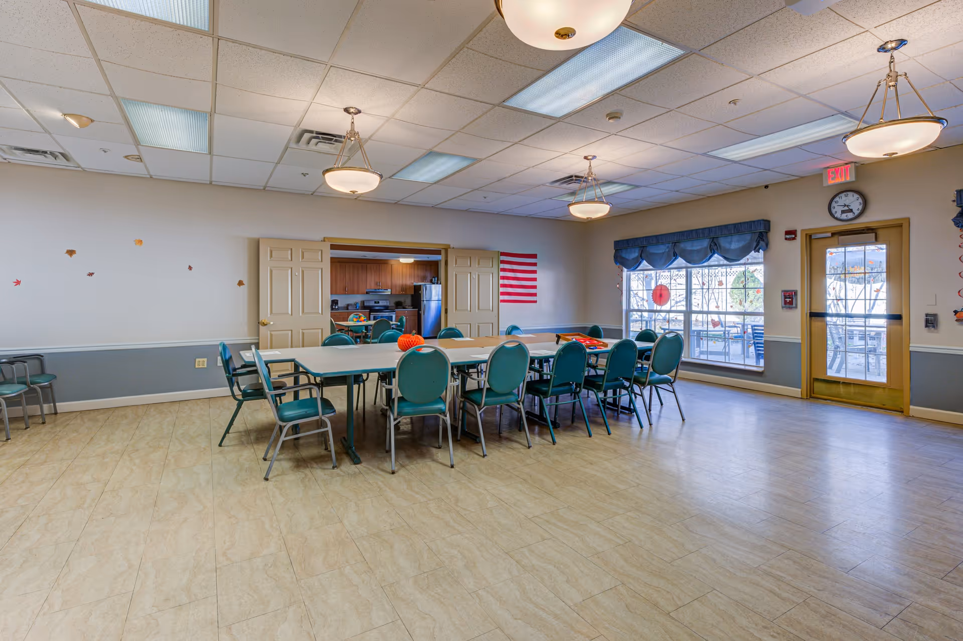 A spacious dining room with a large rectangular table surrounded by green chairs. The room has beige tiled flooring, light-colored walls with a blue-gray wainscoting, and ceiling lights. There is a window with blue valance curtains and a door leading outside. An American flag is displayed on the wall near the entrance to a kitchen area visible through double doors.