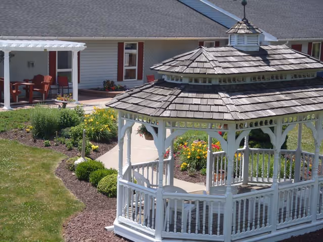 A white wooden gazebo with a shingled roof in a garden area with green grass, bushes, and colorful flowers. In the background, there is a building with white siding, red window shutters, and a covered patio with red chairs and a table.