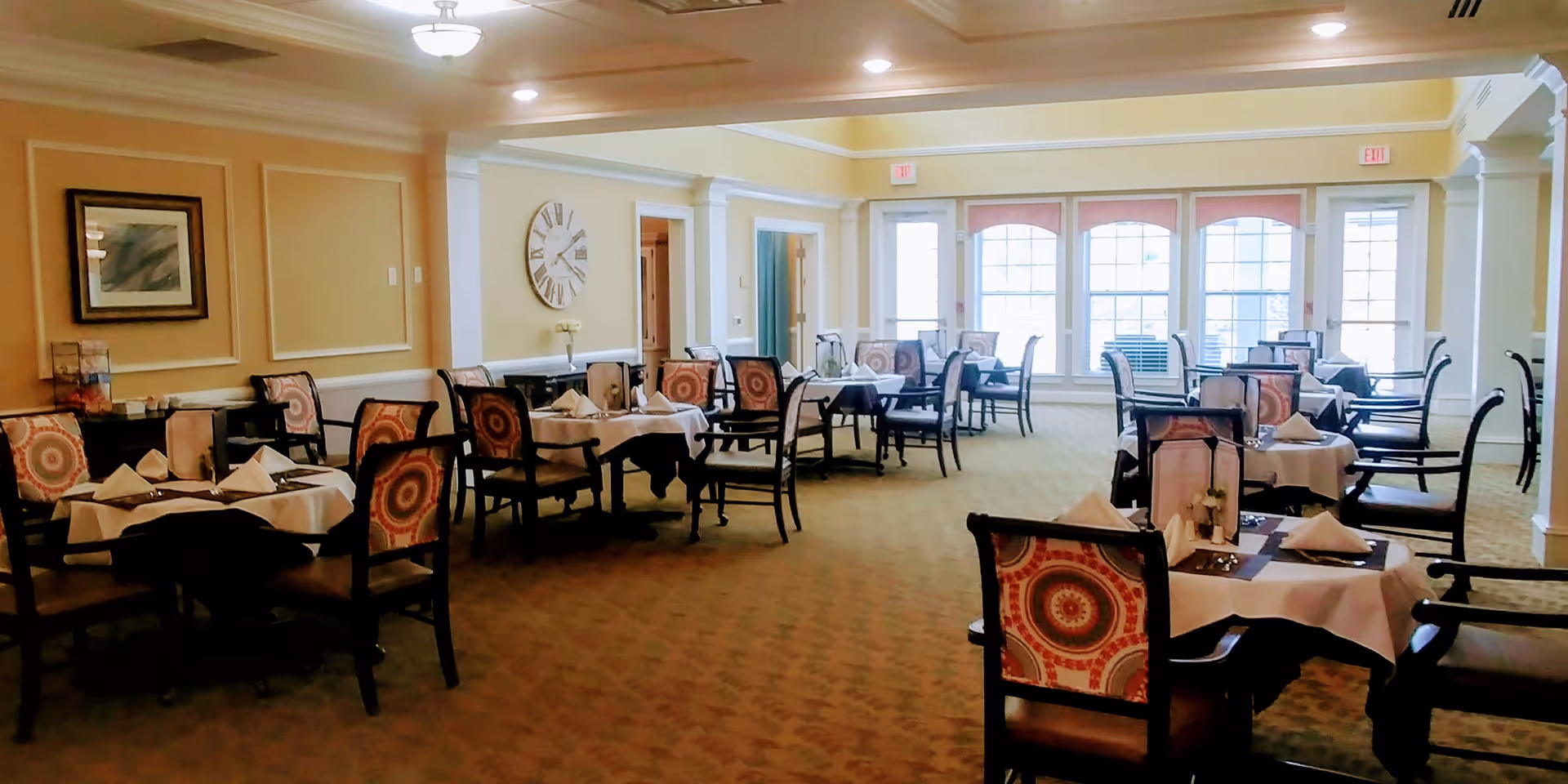 A bright dining room with multiple tables covered in white tablecloths, each set with napkins and menus. The chairs have patterned upholstery with circular designs. Large windows along the back wall let in natural light, and the walls are painted a soft yellow with white trim. A large decorative clock and framed artwork are visible on the walls.