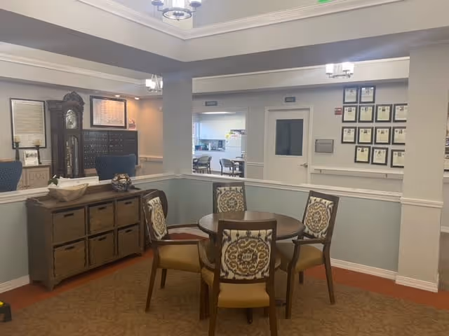 Interior view of a senior living facility common area with a round wooden table surrounded by four upholstered chairs with patterned backs. There is a wooden sideboard with baskets and decorative items on top. The walls are painted light blue with white trim, and framed certificates or documents are displayed on the far wall. A grandfather clock and additional seating are visible in the background.