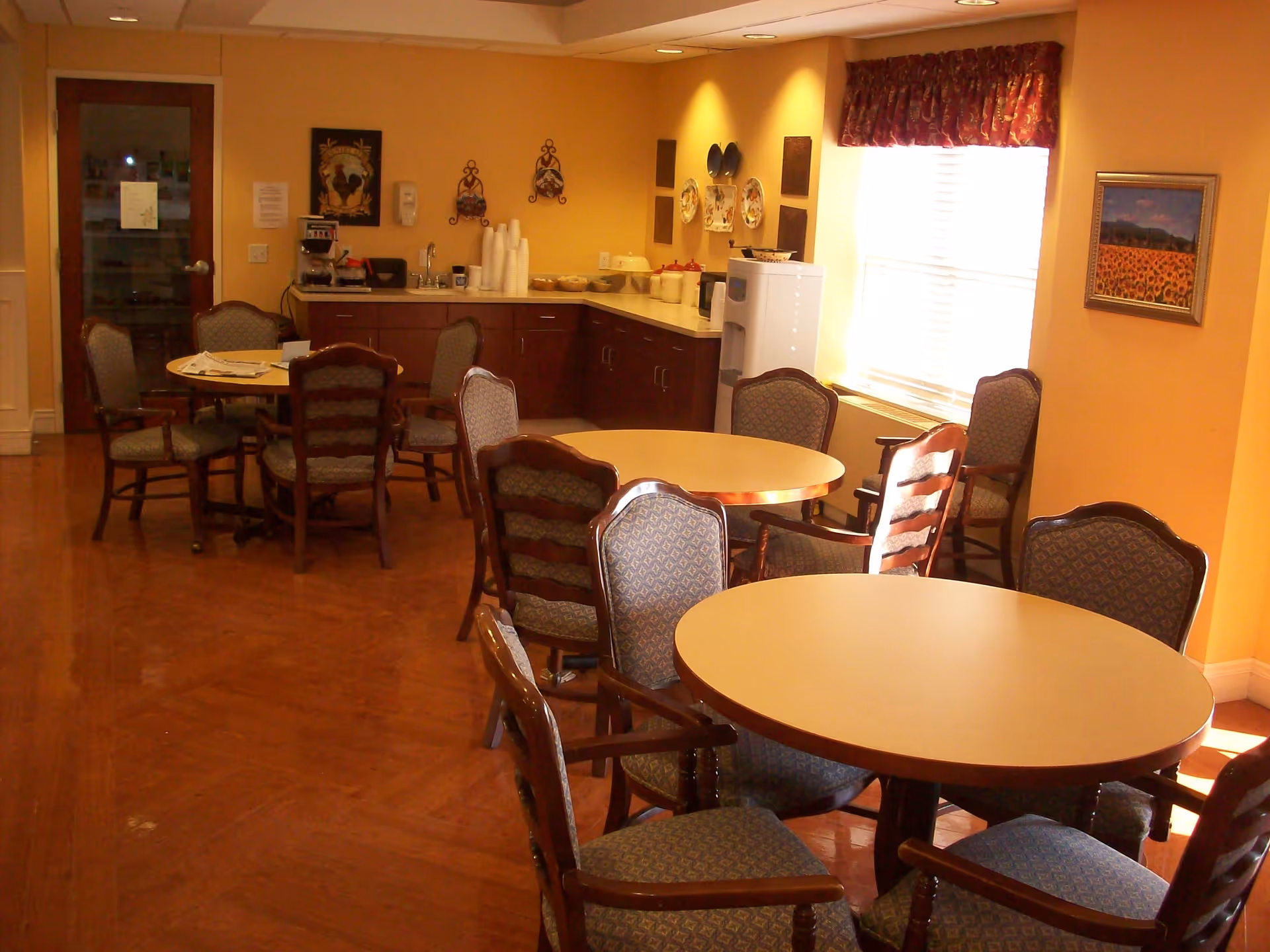 A cozy dining area in a senior living facility with round tables surrounded by cushioned wooden chairs. The room has warm yellow walls, a window with a red valance, and a painting of sunflowers on the wall. There is a counter with a coffee maker, cups, and other kitchen items in the background.