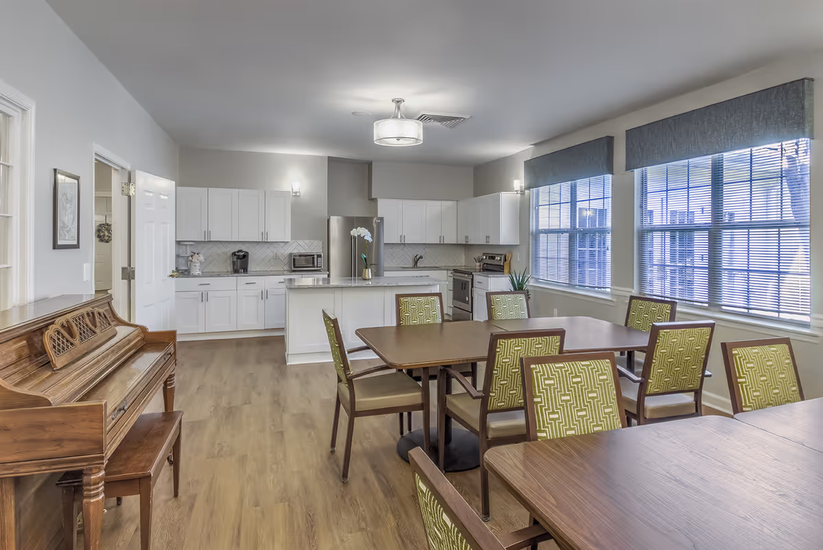 A bright and spacious room featuring a kitchen area with white cabinets, a stainless steel refrigerator, stove, and microwave. In front of the kitchen, there are two wooden dining tables surrounded by chairs with green patterned upholstery. To the left, there is a wooden piano with a matching bench. Large windows with blinds allow natural light to fill the room, and the floor is covered with light wood laminate.