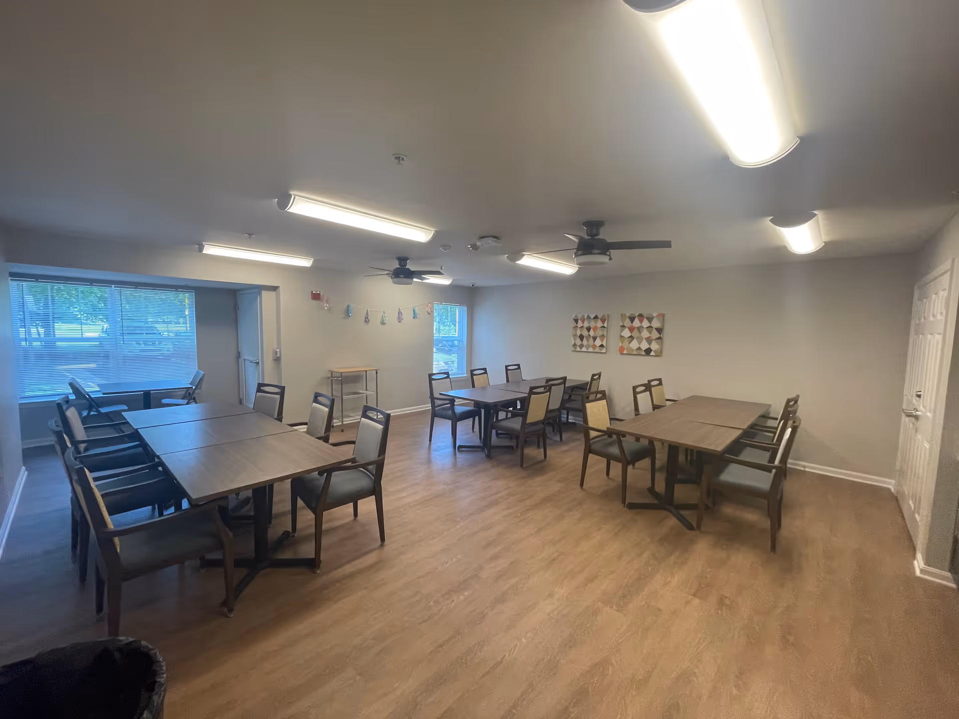 A well-lit room with multiple tables and chairs arranged for group seating, featuring wood flooring, ceiling fans, and wall decorations including two framed artworks and a string of small flags. Large windows allow natural light to enter the space.