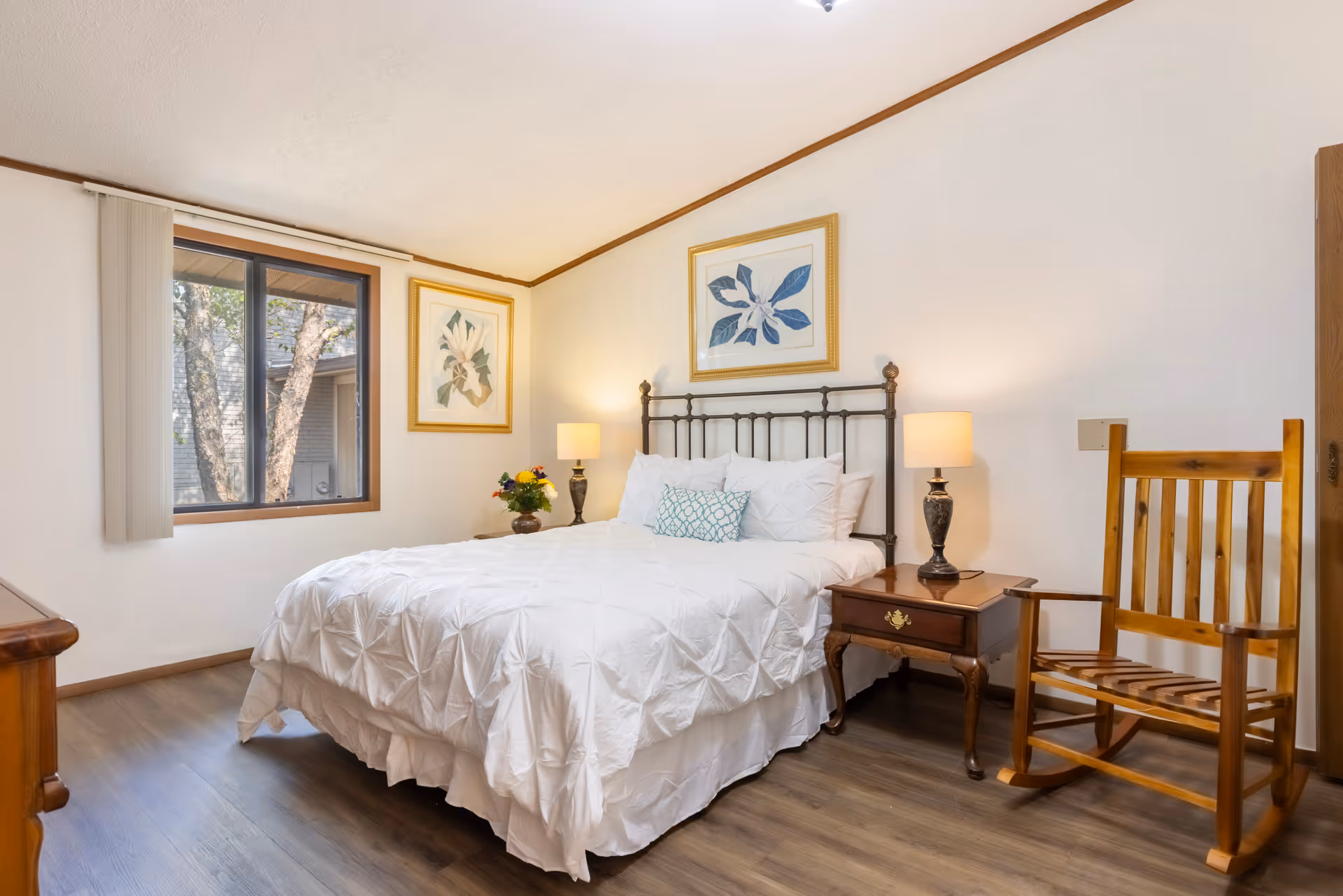 A cozy bedroom with a white bedspread on a metal frame bed, two bedside tables with lamps, a wooden rocking chair, two framed botanical prints on the walls, and a window showing trees outside.