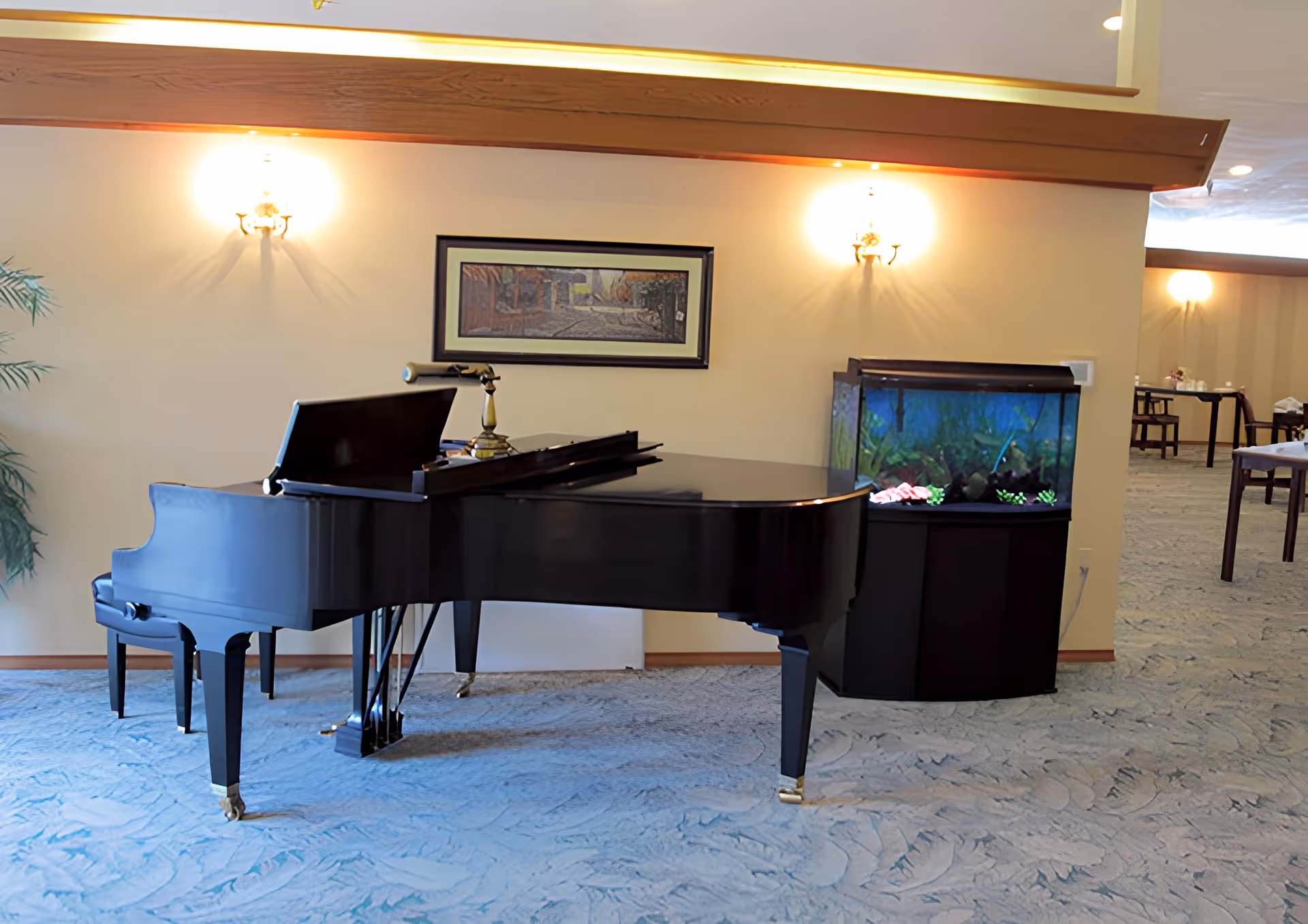 Interior view of a senior living facility lounge area featuring a black grand piano with a piano bench, a fish tank with colorful decorations, wall-mounted lights, a framed painting, and tables with chairs in the background.