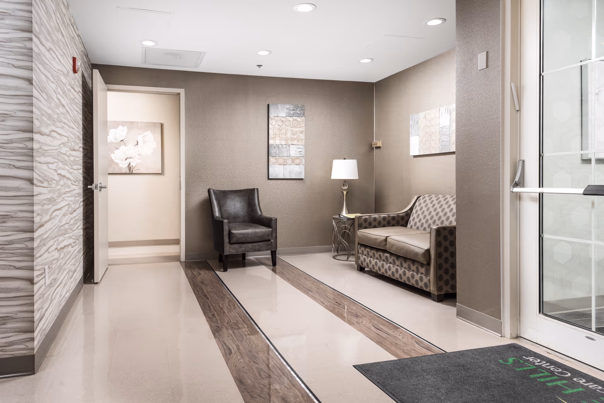 A clean and modern waiting area with a patterned loveseat, a dark leather armchair, a side table with a lamp, and abstract wall art. The floor has a polished finish with wood-like inlays, and a glass door is visible on the right side.