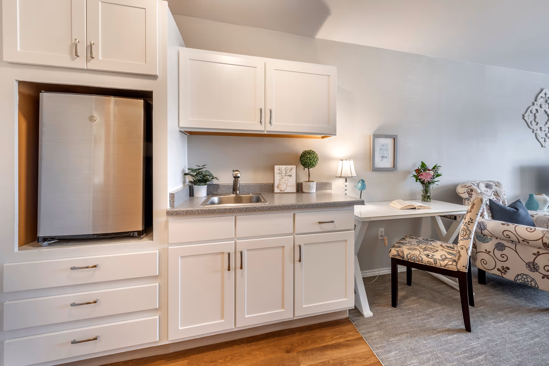 A small kitchenette area with white cabinets, a stainless steel mini refrigerator, a sink, and a countertop decorated with small plants and a framed picture. Adjacent to the kitchenette is a white desk with a floral-patterned chair, a small lamp, a vase with flowers, and an open book. In the background, there is a floral-patterned armchair and a light gray wall with decorative wall art.