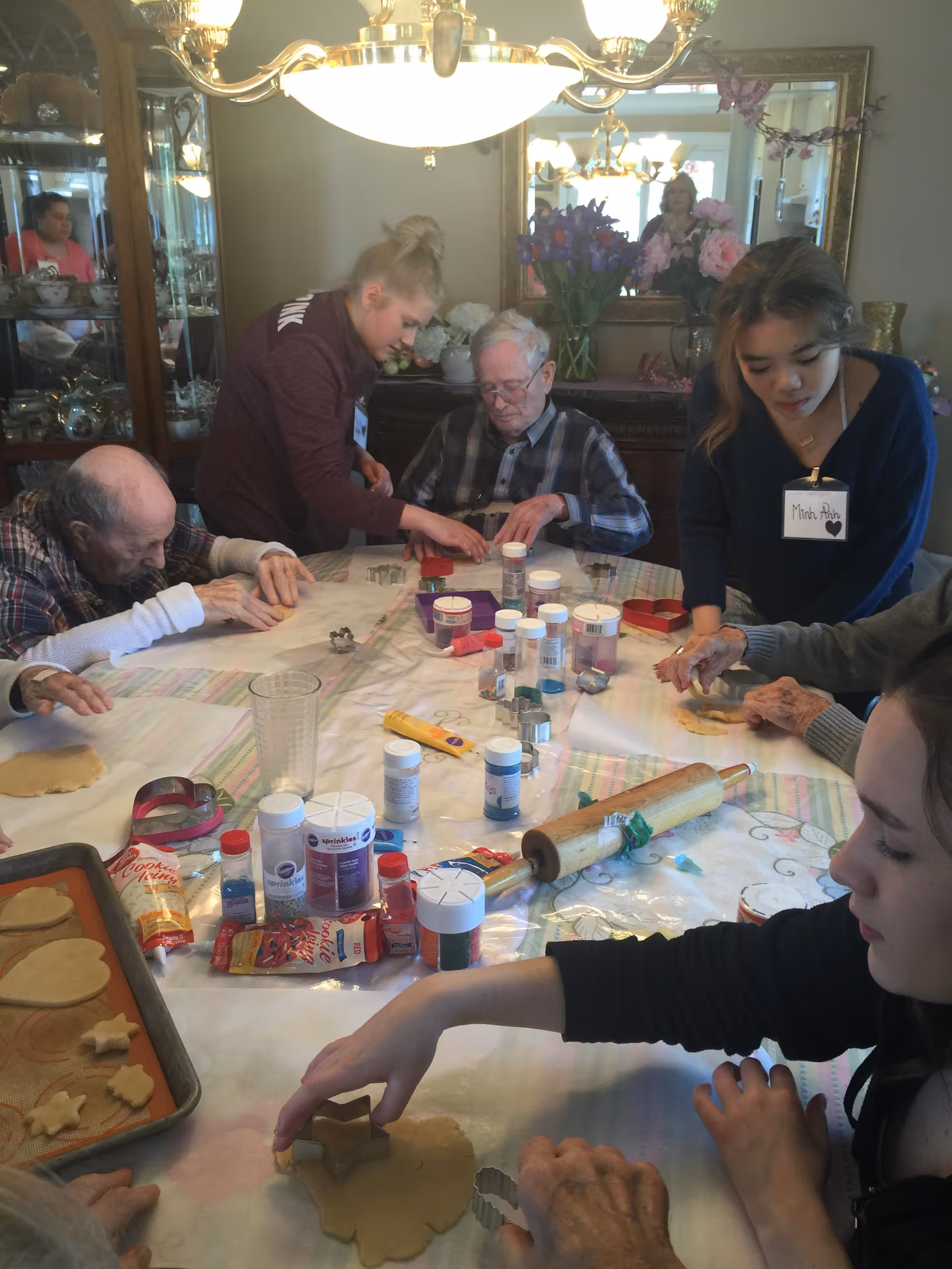 A group of elderly people and caregivers gathered around a table in a dining room, engaged in a cookie decorating activity with various sprinkles, cookie cutters, and dough on the table.