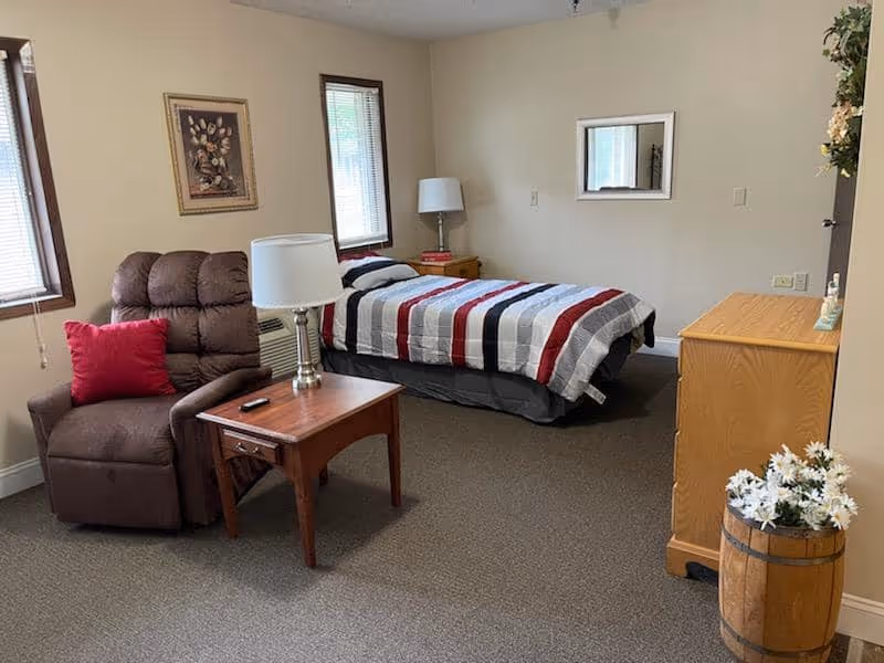 A senior living community bedroom featuring a single bed with a striped red, white, and gray comforter, a brown recliner chair with a red pillow, a wooden side table with a lamp, a wooden dresser, two windows with blinds, a framed picture on the wall, a mirror, and a decorative barrel with white flowers.