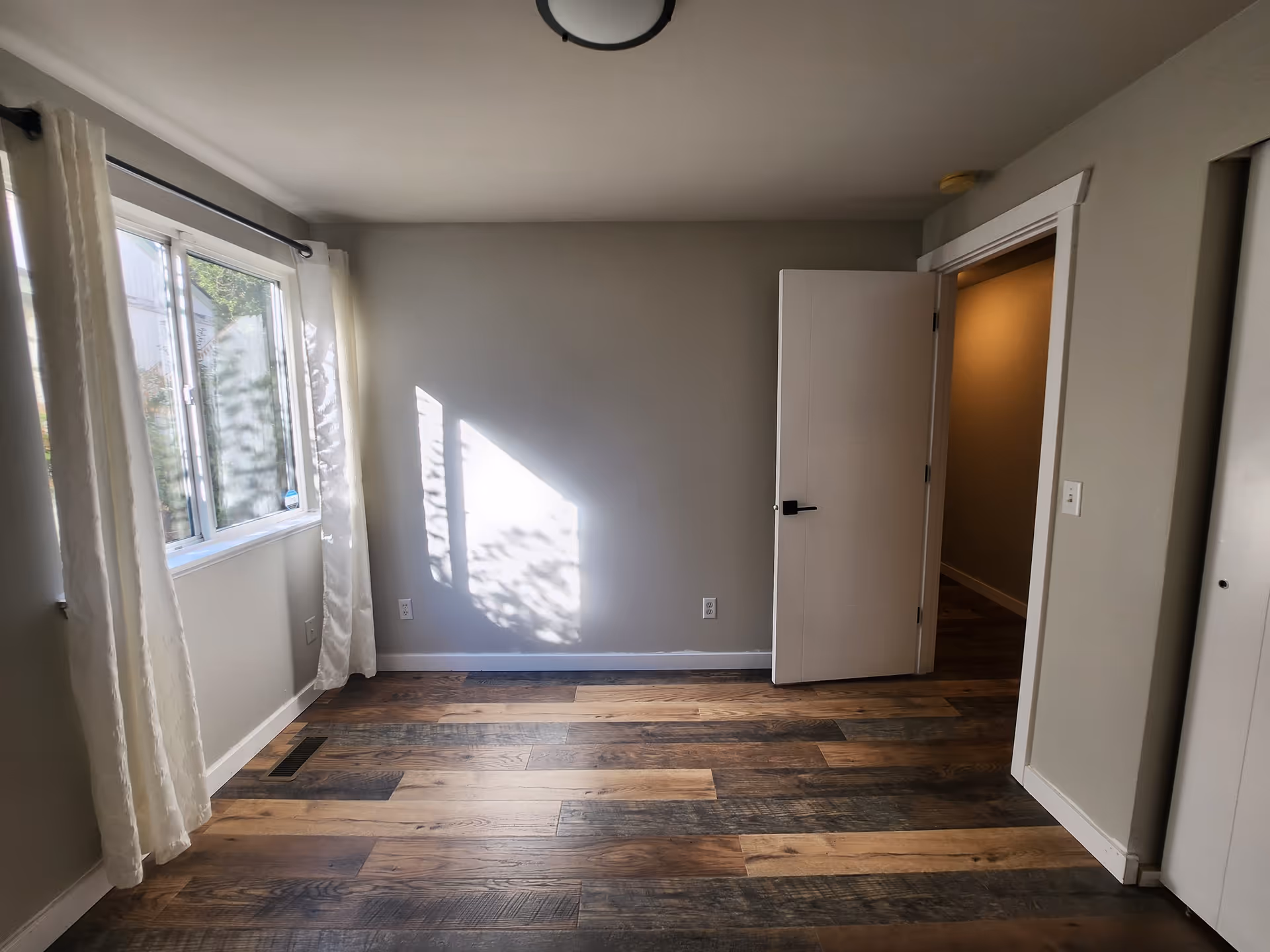 Empty room with wooden flooring, light gray walls, a window with white curtains on the left, and a white door partially open on the right leading to a hallway.