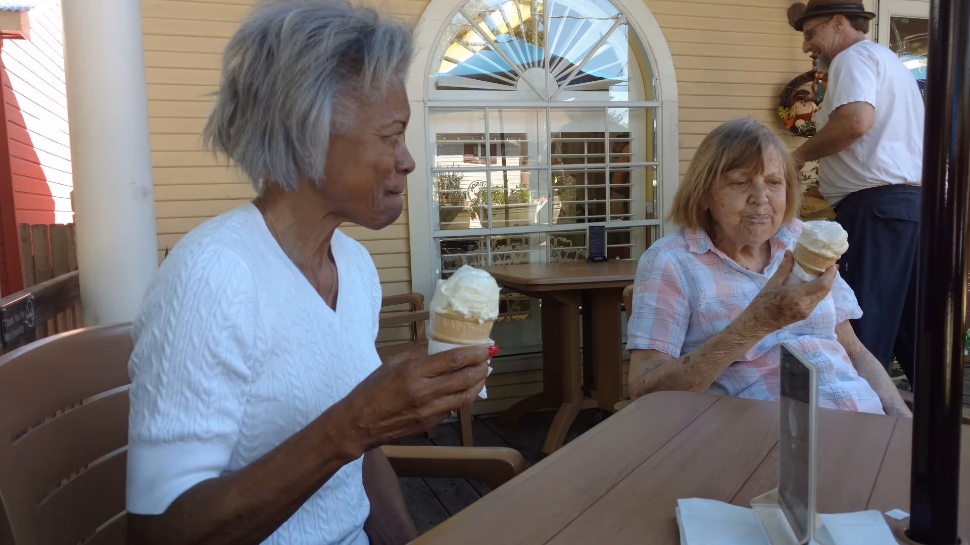Two elderly women sitting at a wooden outdoor table enjoying ice cream cones. One woman is wearing a white sweater and the other a plaid shirt. A man in a white shirt and hat is standing in the background near a yellow building with a window.