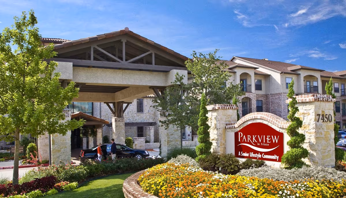 Exterior view of Parkview in Frisco senior living community showing the entrance with a covered drop-off area, stone pillars, landscaped garden with colorful flowers, and a large red sign displaying the community name and slogan.