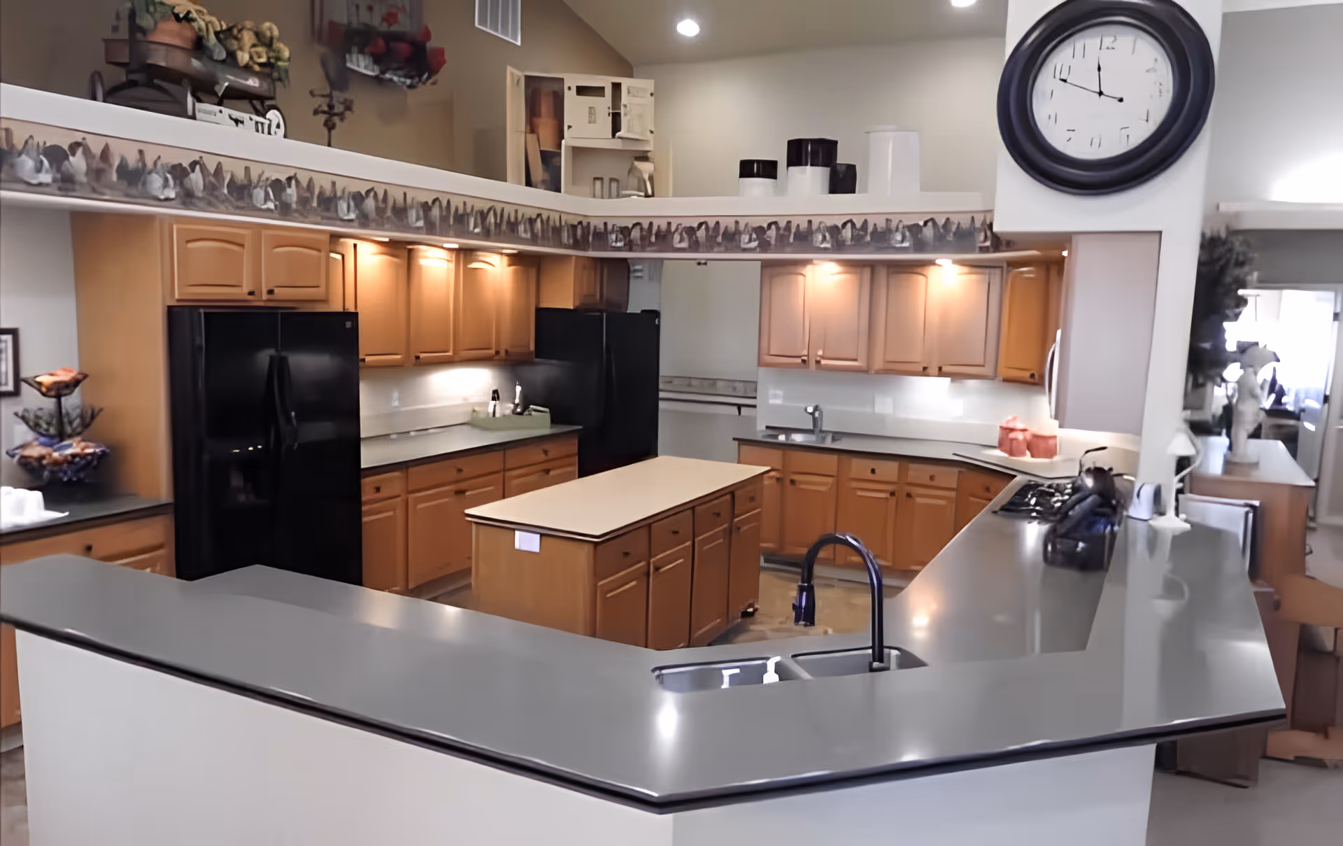 Open communal kitchen with a central island, wraparound countertops, wooden cabinets, black appliances and a large wall clock.