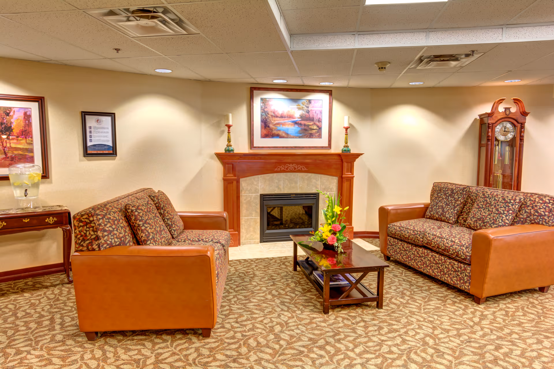 Cozy retirement community lounge with two patterned sofas facing a fireplace, a coffee table with flowers, and a grandfather clock.