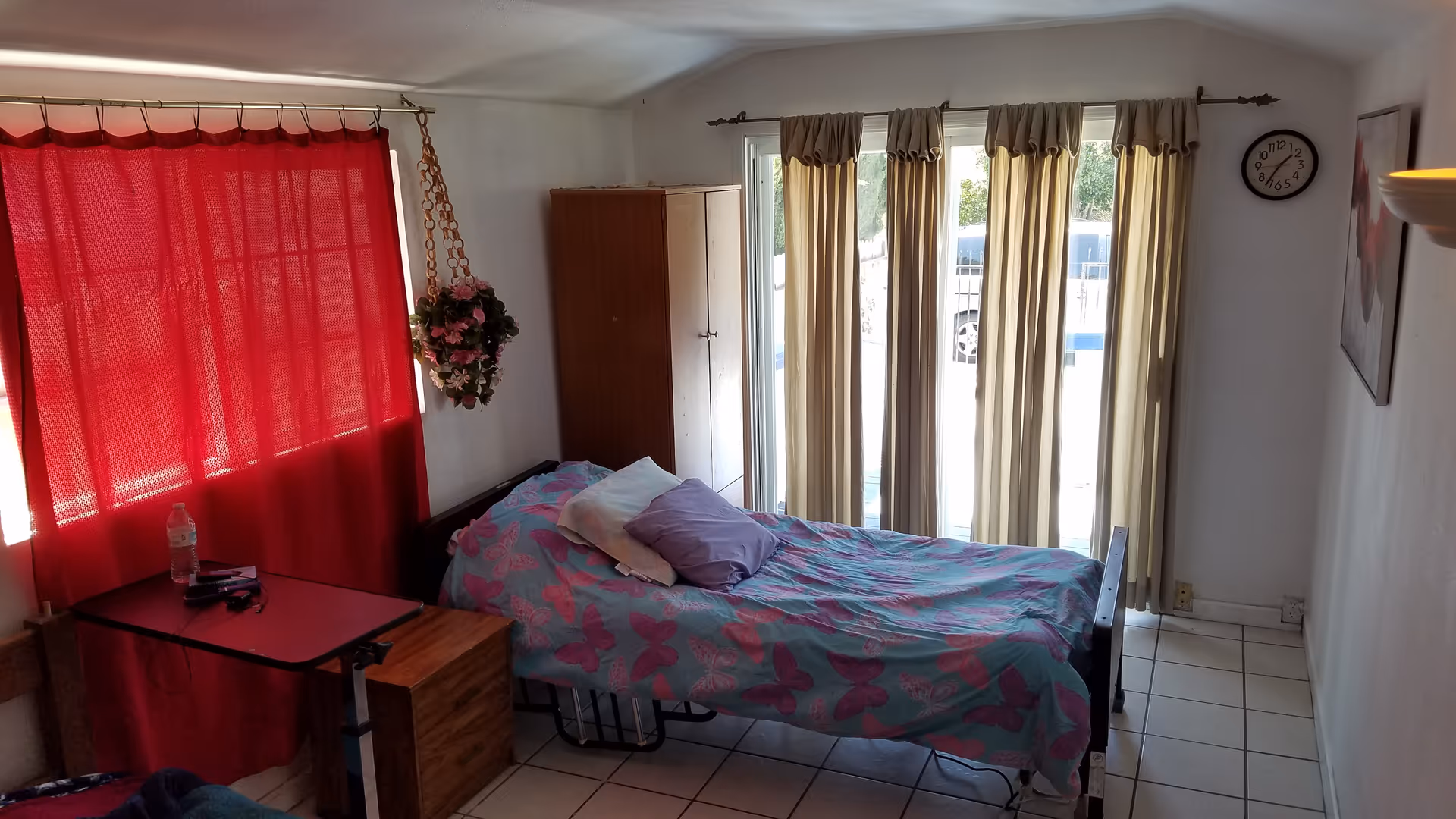 A small bedroom with a single bed covered in a butterfly-patterned blanket and two pillows. There is a red curtain covering a window on the left side, a wooden cabinet in the corner, and beige curtains covering a glass door or window on the right side. A clock is mounted on the wall above the curtains, and a hanging flower pot is near the bed.