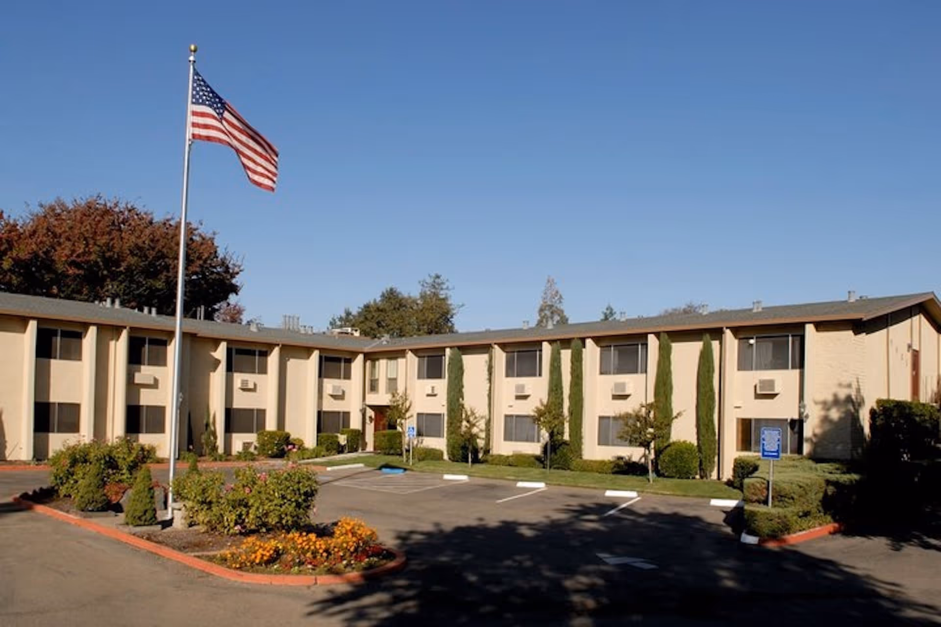 Exterior view of a two-story senior living facility building with beige walls and multiple windows. There is a parking lot in front with marked spaces, some greenery including bushes and small trees, and an American flag on a flagpole near the center-left of the image under a clear blue sky.