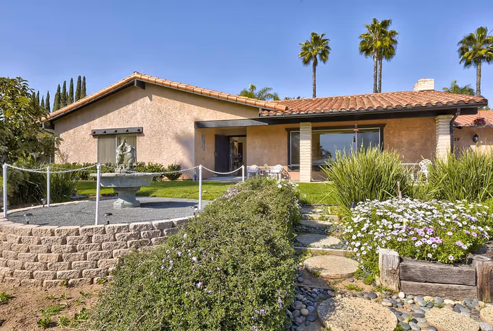 Exterior view of a single-story building with a tiled roof, surrounded by a well-maintained garden featuring a stone fountain, green shrubs, flowering plants, and palm trees under a clear blue sky.