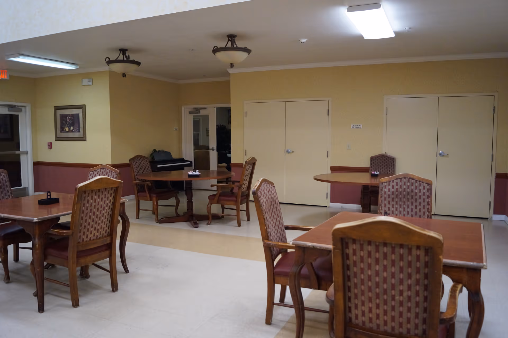 A dining area with several wooden tables and cushioned chairs arranged around the room. The walls are painted yellow with a maroon wainscot. There is a piano in the corner near a door, and two sets of double doors are visible on the far wall. Ceiling lights illuminate the space.
