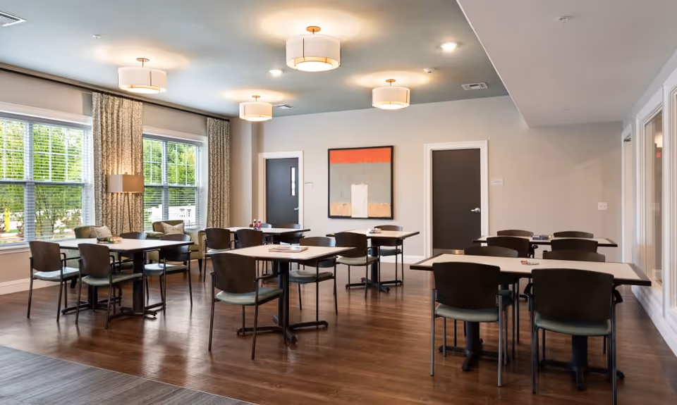Bright communal dining room with multiple tables and chairs, large windows, modern light fixtures, and wall art.