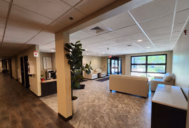 Interior view of a senior living facility lobby area with beige sofas and armchairs arranged near large windows letting in natural light. A potted plant is placed near a column, and a reception desk is visible to the left. The floor has a marbled pattern, and the ceiling has recessed lighting and ventilation panels.