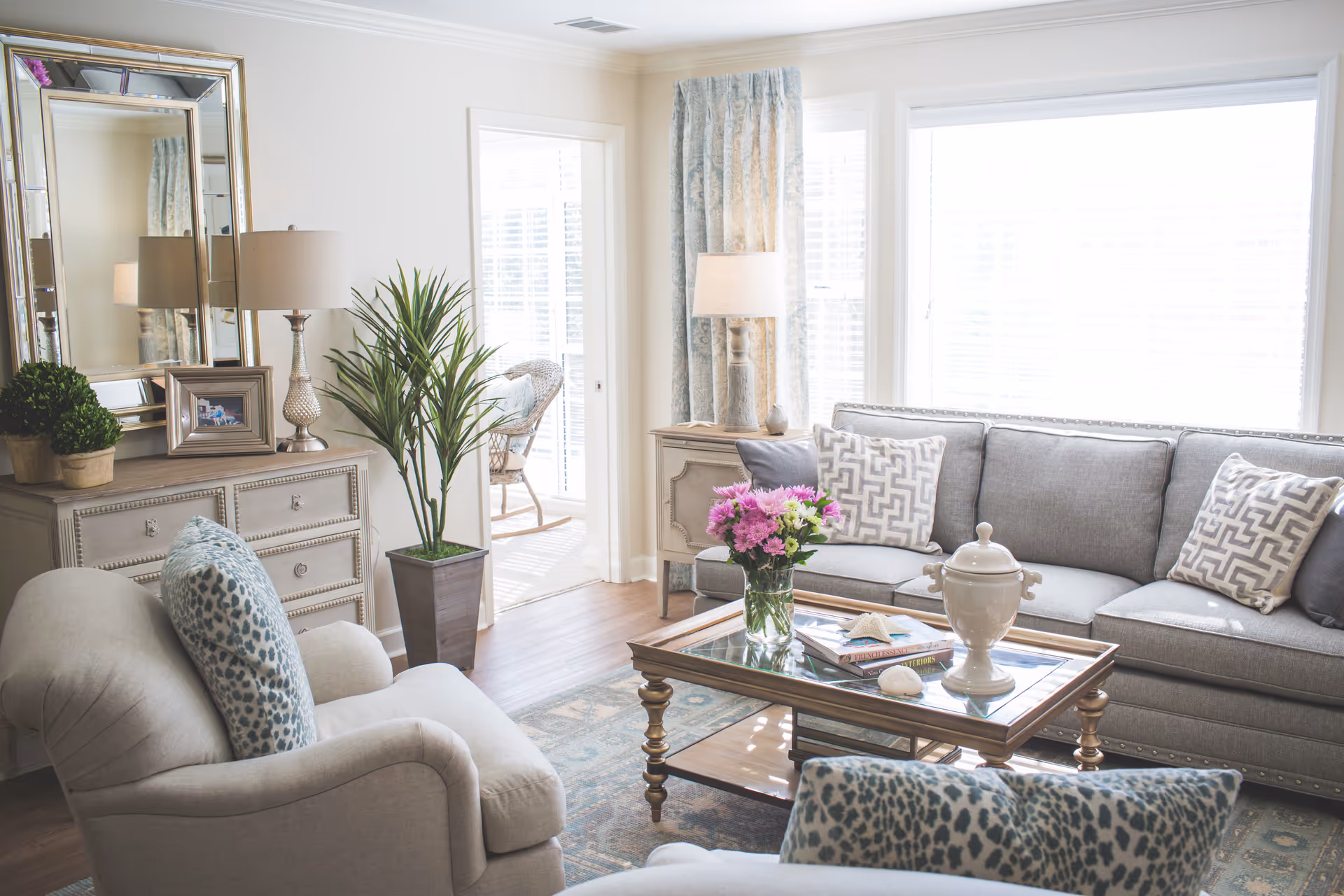 Bright, elegantly decorated living room with a gray sofa, upholstered armchairs, a glass-top coffee table with flowers, and large sunlit windows.