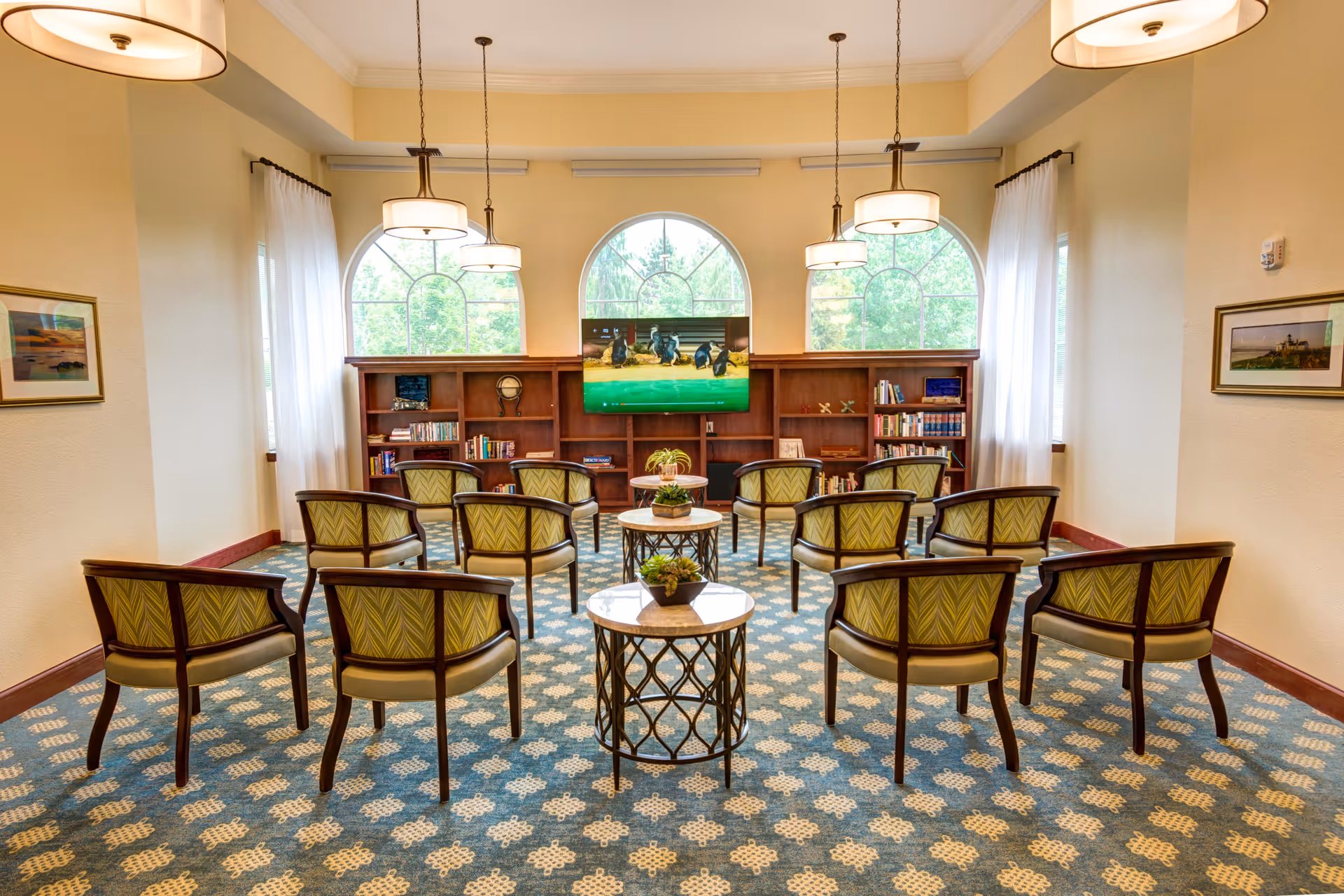 A bright and spacious senior living common area with multiple green patterned chairs arranged in rows facing a wall-mounted flat-screen TV. The room features large arched windows with white curtains, wooden bookshelves filled with books and decorative items, three round tables with small plants, and hanging pendant lights from the ceiling. The carpet has a blue and beige geometric pattern, and framed artwork is hung on the cream-colored walls.