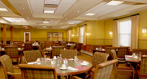 A spacious dining room with multiple round tables set with cups, glasses, and napkins. The room has cushioned chairs with plaid upholstery, beige walls with decorative wallpaper, and large windows with curtains allowing natural light. Ceiling lights illuminate the room.