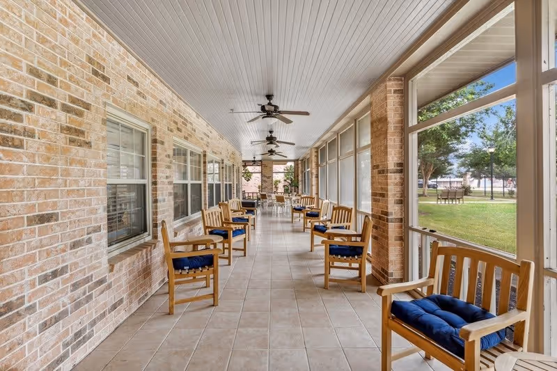 A long covered porch area with multiple wooden chairs featuring blue cushions arranged along both sides. The porch has a tiled floor, brick walls, ceiling fans, and large windows looking out onto a grassy outdoor area with trees and additional seating.