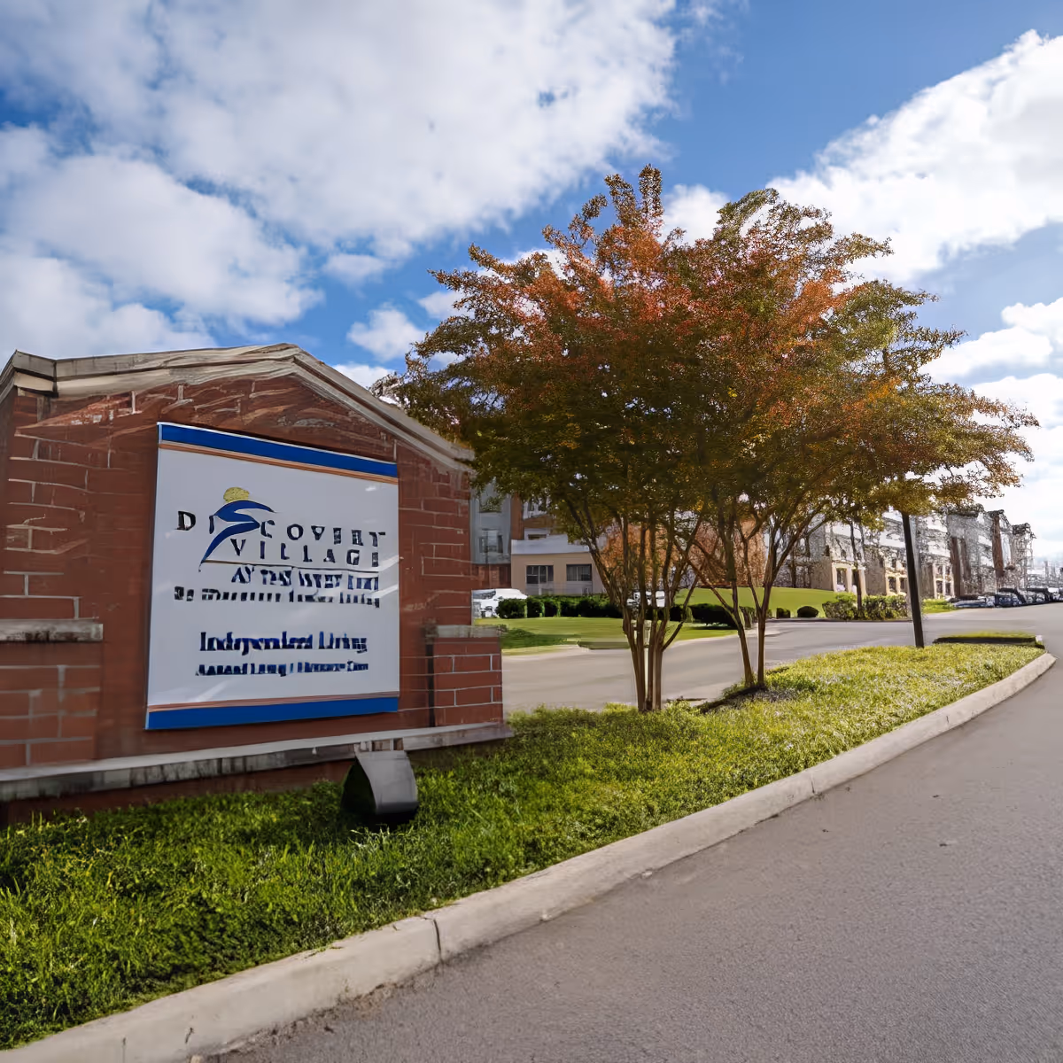 Exterior view of Discovery Village At the West End facility showing a brick sign with the facility name, a tree with autumn-colored leaves, a driveway, and part of the building under a partly cloudy sky.