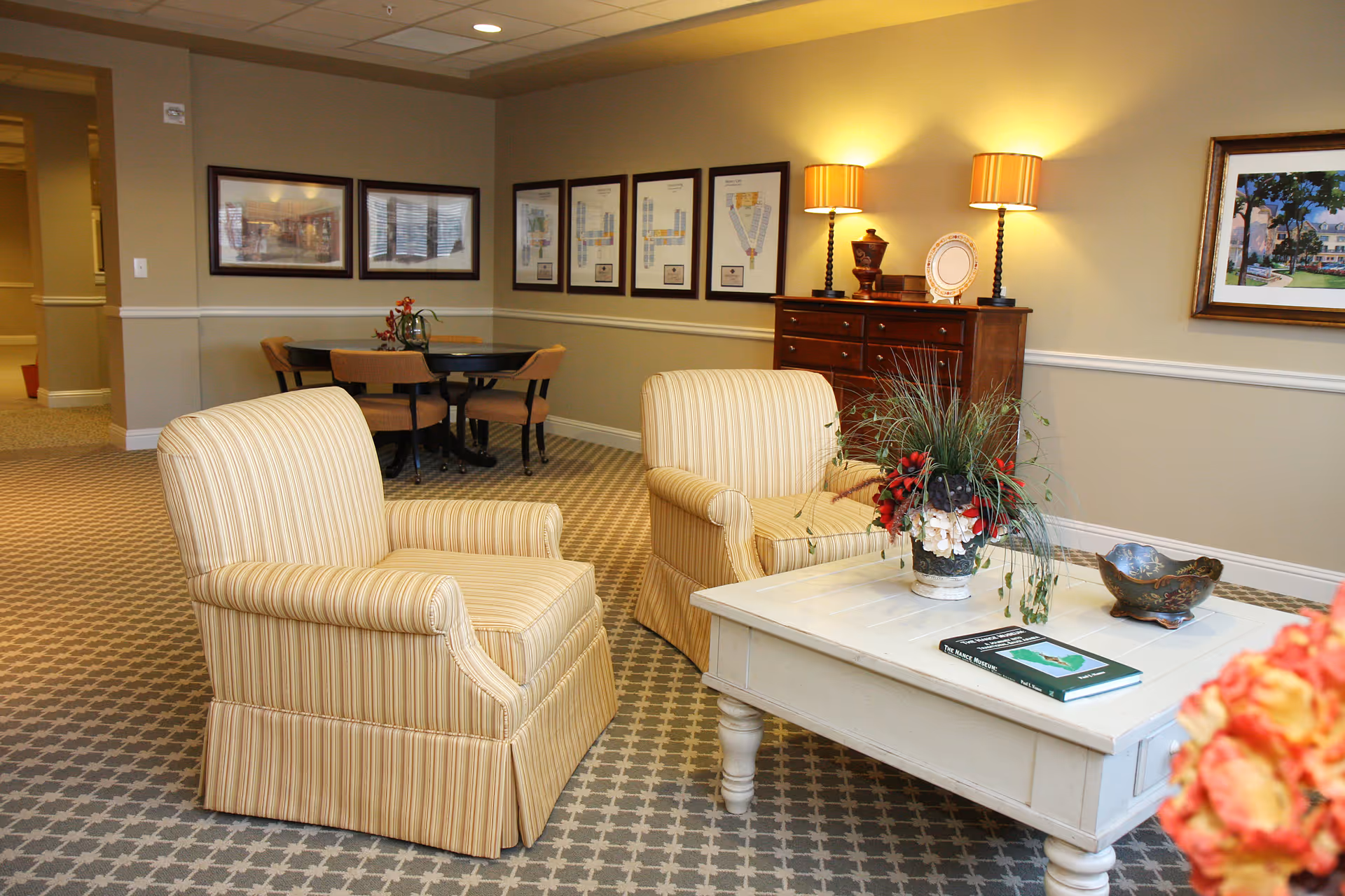 A cozy sitting area in a senior living facility with two striped armchairs facing a white coffee table adorned with a floral arrangement and a book. In the background, there is a wooden dresser with two table lamps and decorative items, framed pictures on the walls, and a round table with four chairs.