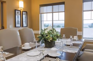 A well-lit dining room with a long wooden table set for a meal, featuring white cups, plates, glasses, and silverware. A floral centerpiece is placed in the middle of the table. Large windows with blinds allow natural light to fill the room.