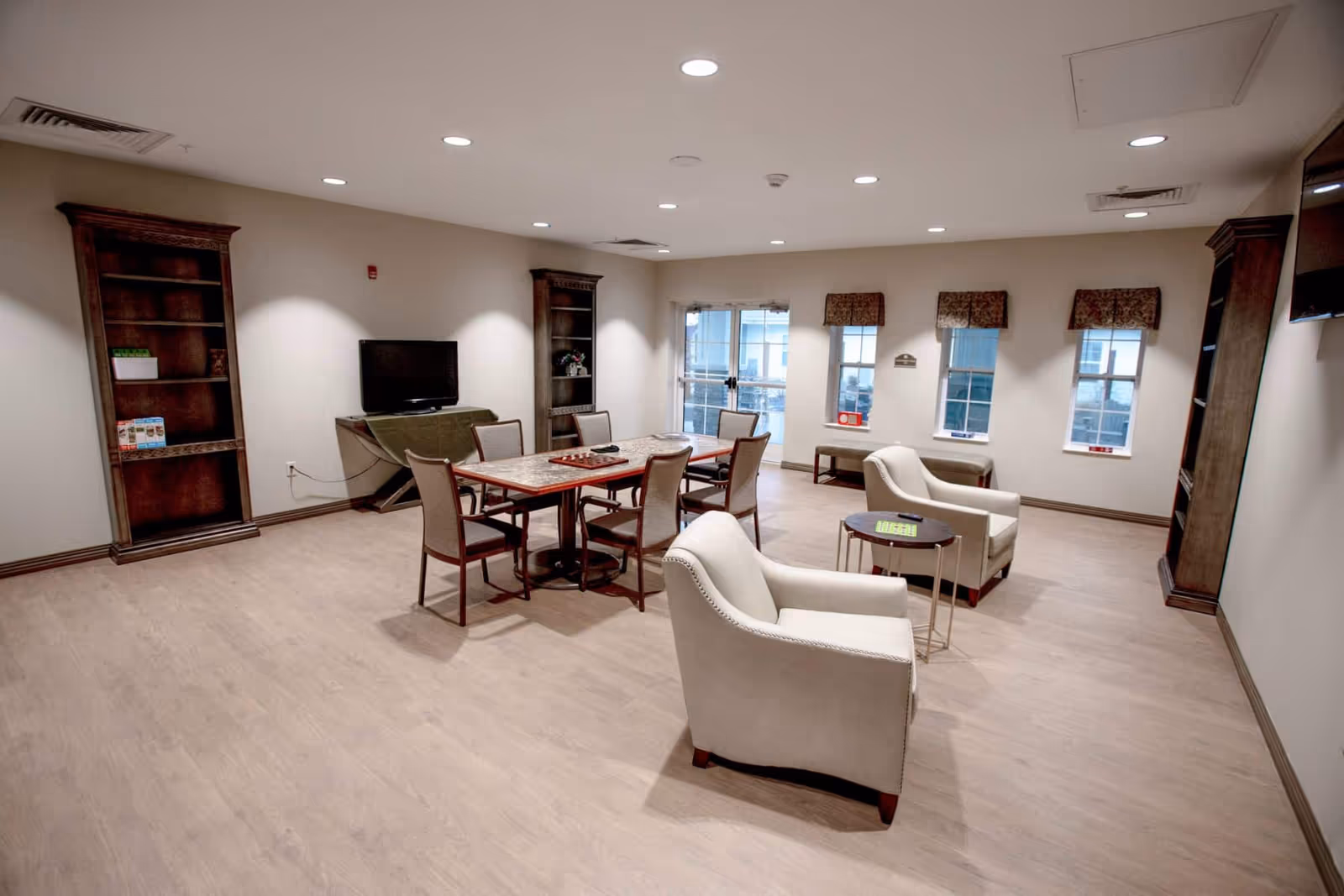 A spacious common area with light wood flooring, beige walls, and recessed ceiling lights. The room features a rectangular table with six chairs, two beige armchairs with a small round table between them, two tall wooden bookshelves, a TV on a stand, and three windows with valances. There is a door with glass panels leading outside.