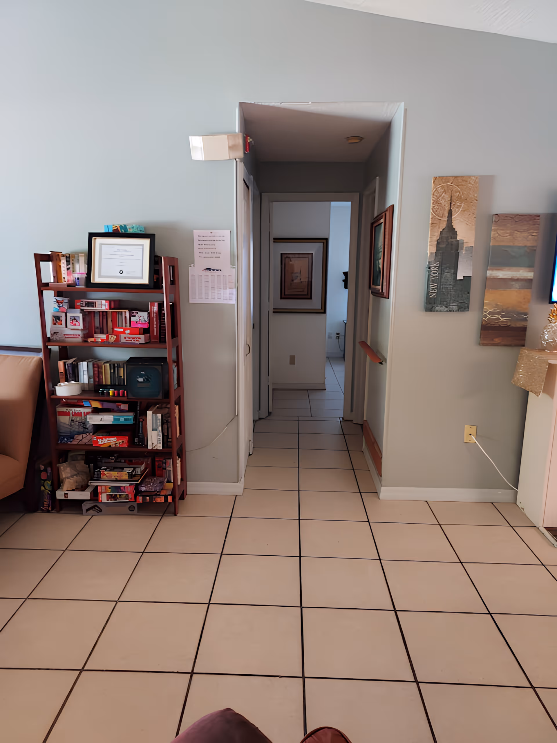 Interior view of a senior living facility hallway with beige tiled floor and light gray walls. On the left side, there is a wooden bookshelf filled with books, framed pictures, and various items. On the right wall, there are three decorative wall hangings, including one with an image of the New York skyline. The hallway leads to another room with framed artwork visible on the walls.